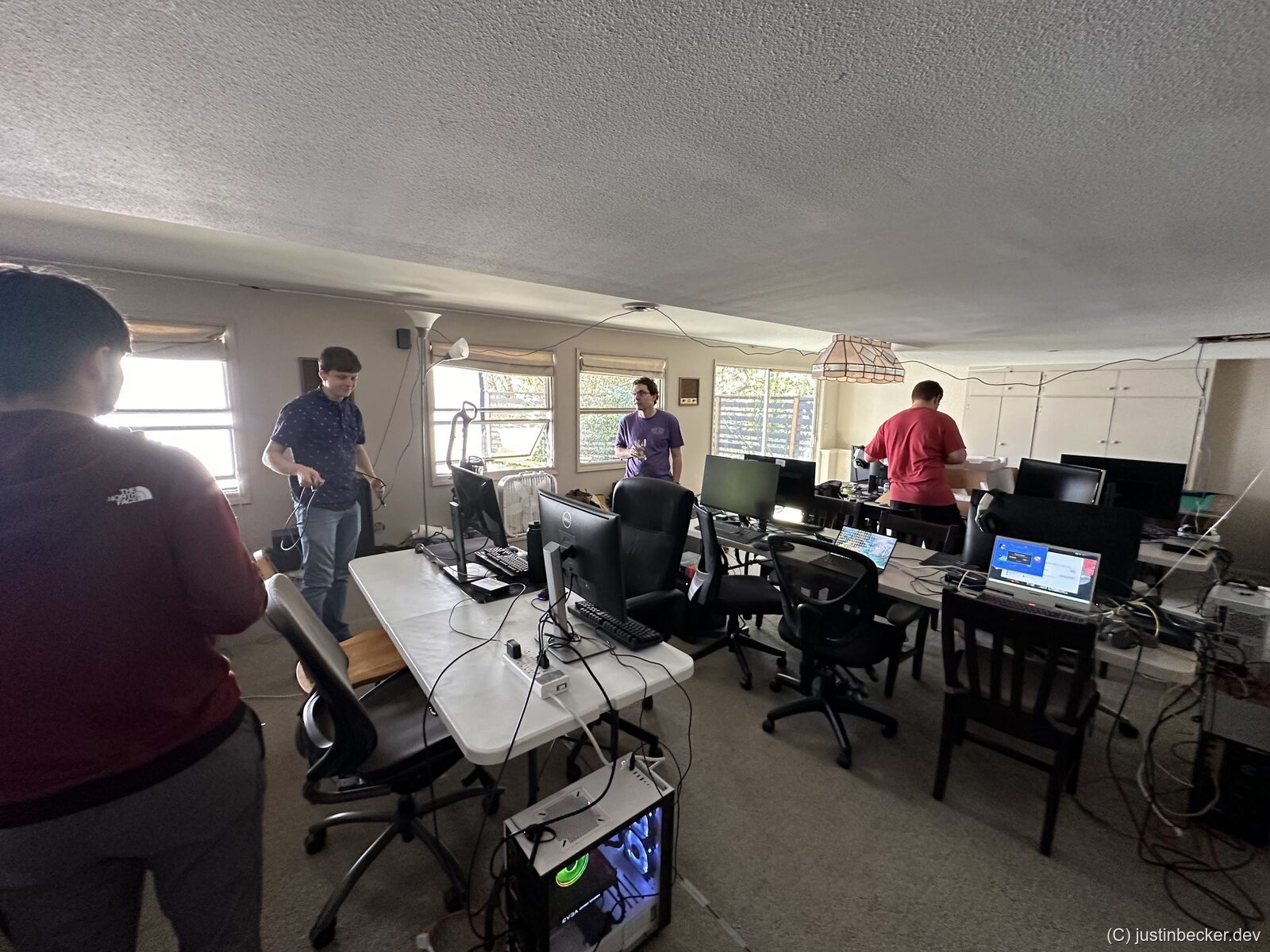 A few people setting up PCs, monitors, and cabling on folding tables in a room before a LAN party begins
