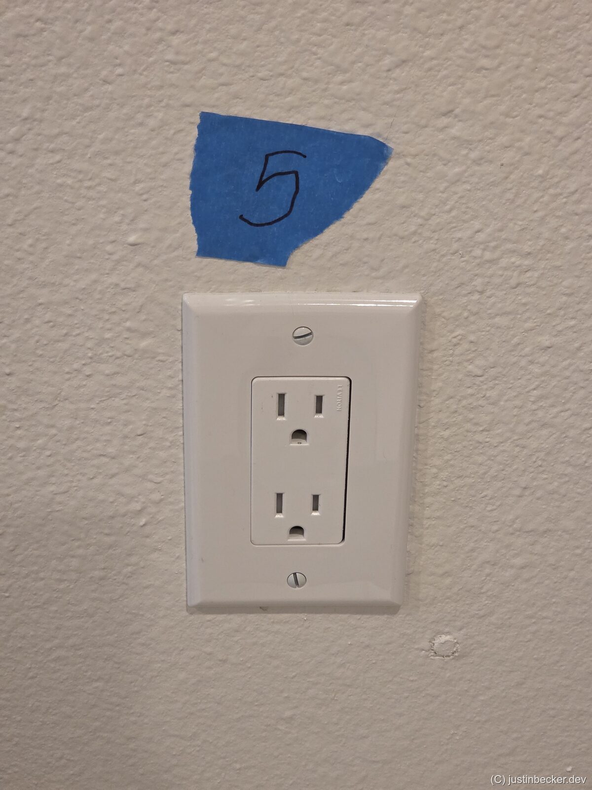 A wall outlet with a piece of blue painter's tape beside it marked with a handwritten 5